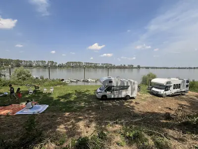 Zwei Wohnmobile am Flussufer mit Picknickdecke und Campern unter blauem Himmel im Fishing Camp Serravalle.